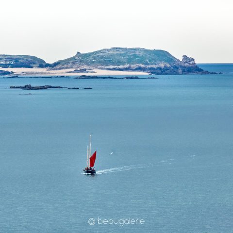 Voilier traditionnel avec voile rouge naviguant devant les côtes rocheuses et les plages de l'île de Cézembre à Saint-Malo.