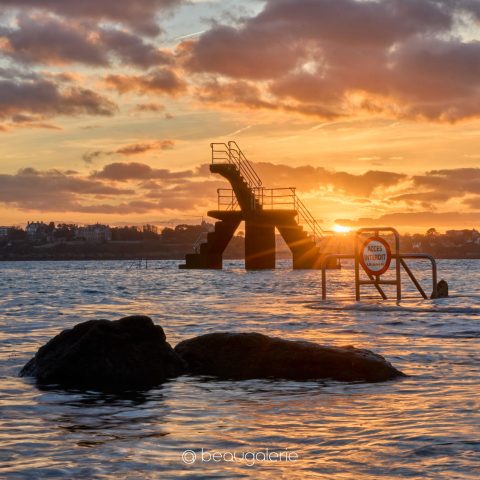 Photographie d'art capturant le Crépuscule Plongeoir Saint Malo à contre-jour avec soleil rayonnant et rochers sombres.