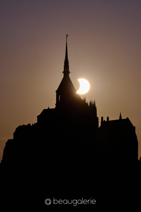 Éclipse solaire du Mont Saint Michel, disque solaire en croissant touchant la flèche gothique en silhouette, ciel violet orangé, format standard, photographie d'art